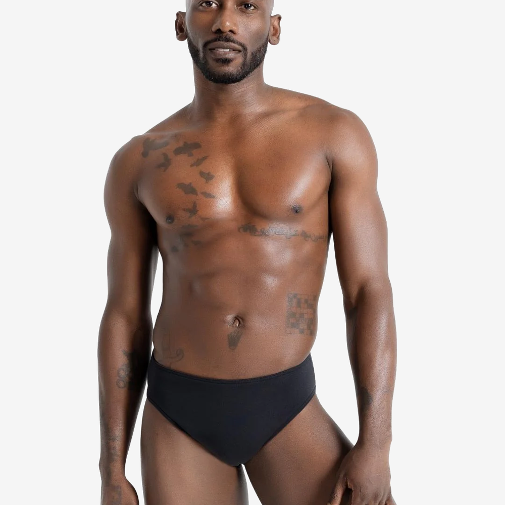A young man with a close-to-face beard and shaved head is standing against a plain light grey background. He is shirtless and wearing a black-colored compression garment. He has tattoos and a neutral expression and is looking directly at the camera.