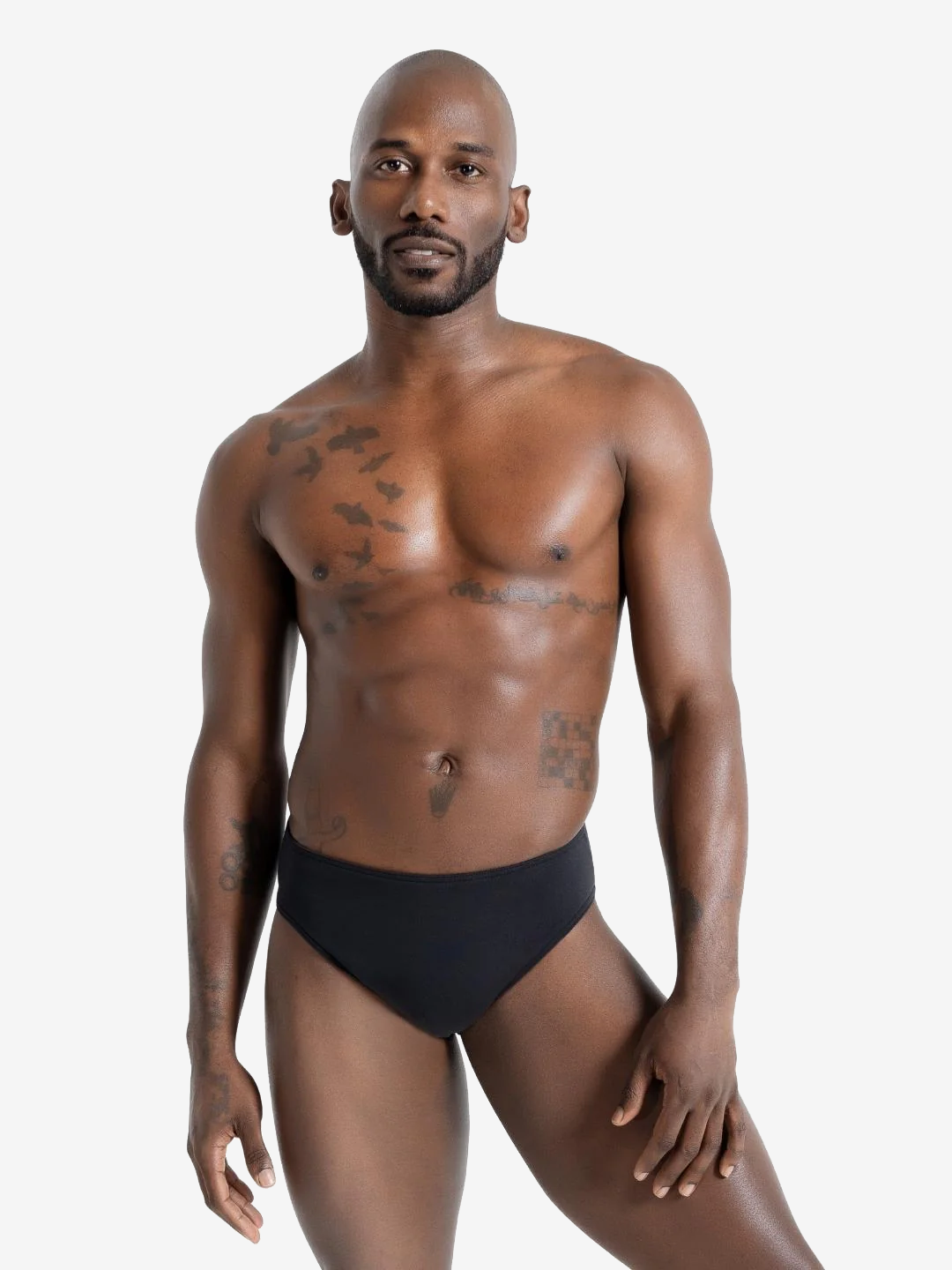 A young man with a close-to-face beard and shaved head is standing against a plain light grey background. He is shirtless and wearing a black-colored compression garment. He has tattoos and a neutral expression and is looking directly at the camera.