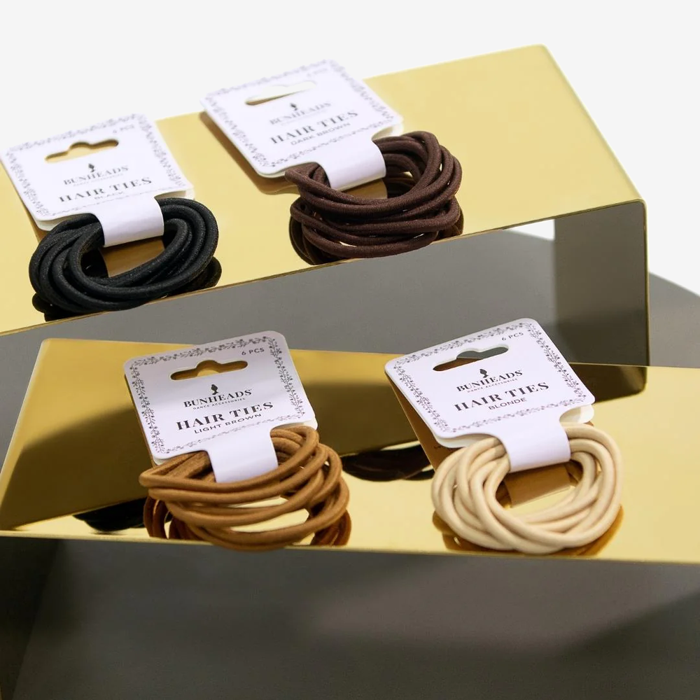 
                      
                        Four packages of various colored hair ties, blonde, light brown, brown, and black, sit on two gold stands.
                      
                    