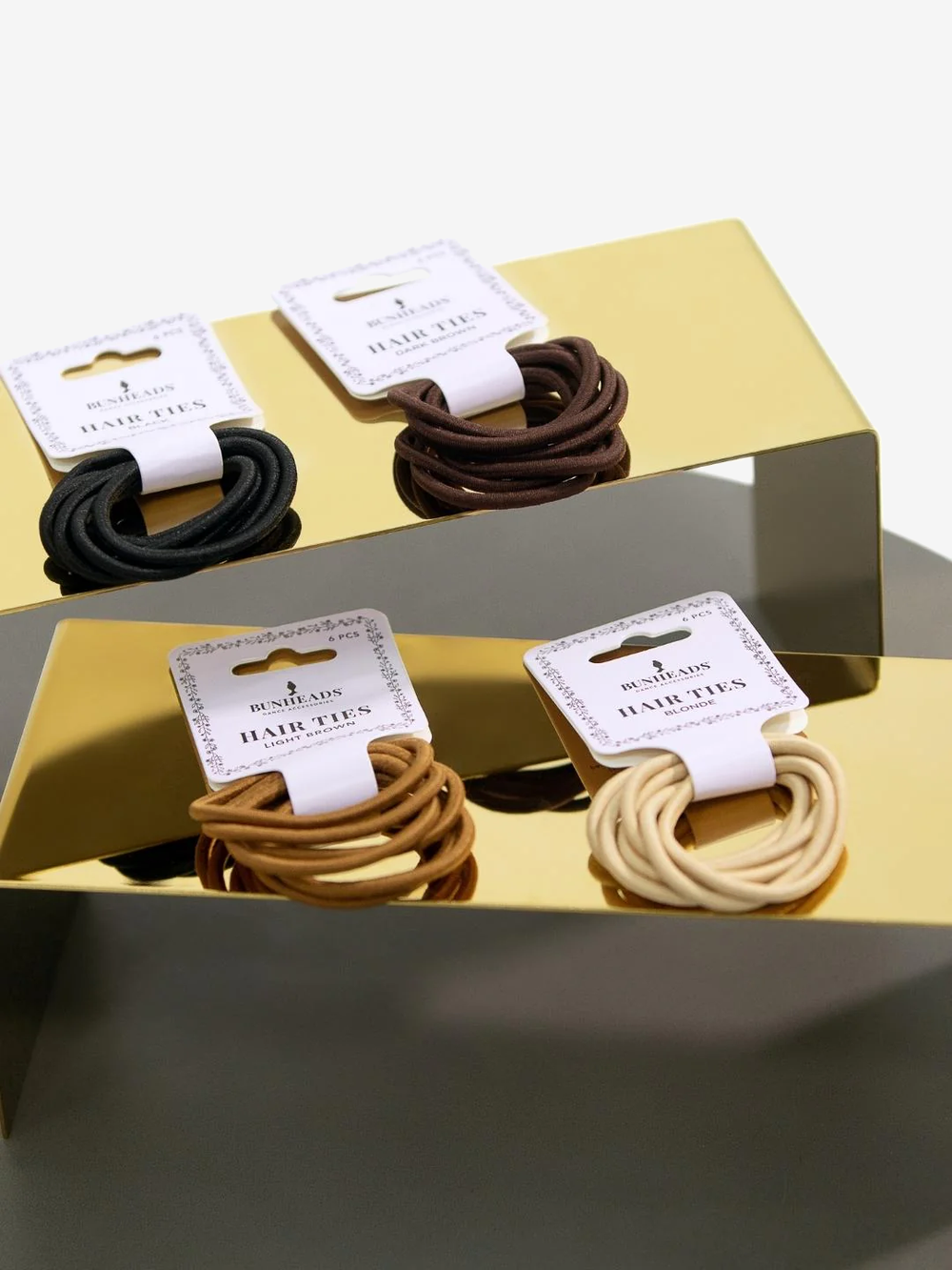 Four packages of various colored hair ties, blonde, light brown, brown, and black, sit on two gold stands.