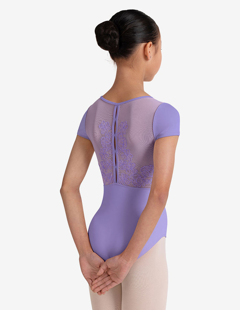 
                      
                        Young girl with a black bun wearing a lilac leotard with mesh details on a white background
                      
                    