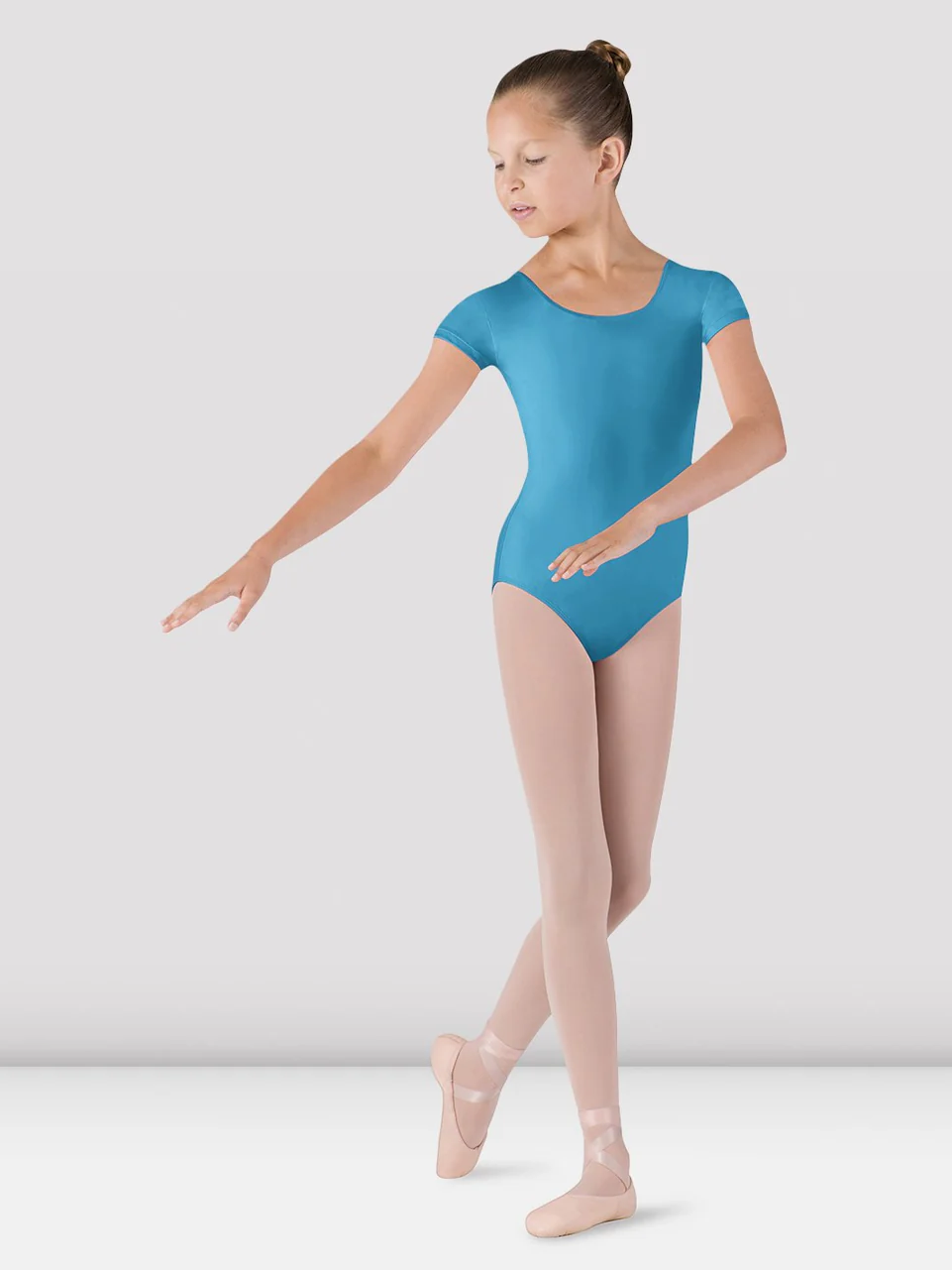 A young ballet dancer stands gracefully in a ballet pose. She is wearing a bright turquoise leotard, light pink tights, and ballet shoes. Her hair is neatly tied back in a bun, and she is positioned against a plain grey background.