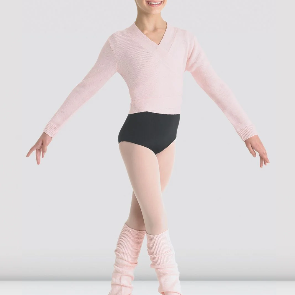 A ballet dancer poses with her arms stretched to the sides. She is wearing a black leotard, pink tights, pink shoes, pink legwarmers, and a knitted light pink ballet wrap sweater. The background is plain light grey.