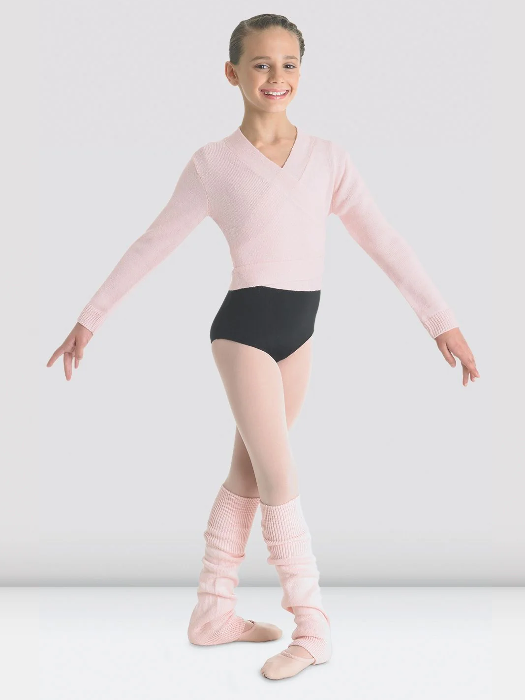 A ballet dancer poses with her arms stretched to the sides. She is wearing a black leotard, pink tights, pink shoes, pink legwarmers, and a knitted light pink ballet wrap sweater. The background is plain light grey.