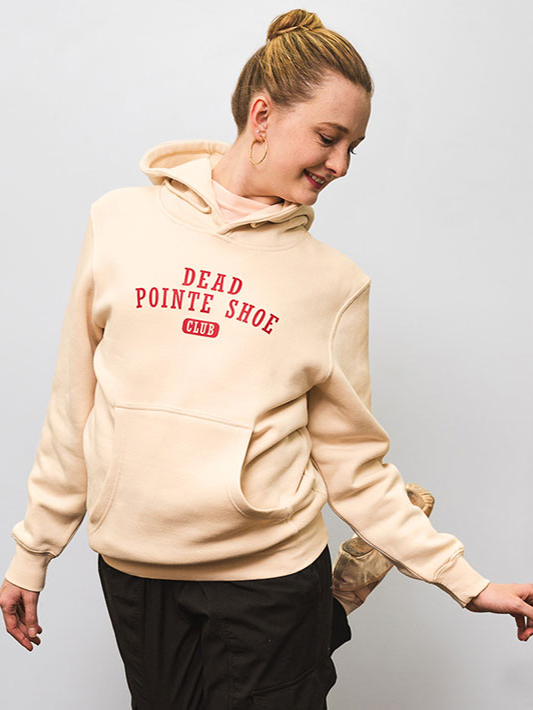 A female with a blond bun looks off to her left, back leg raised behind her. She wears black warm-up pants, pointe shoes, and a cream hoodie with red text, 
