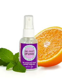 A small spray bottle with a purple DE-FEET DA FUNK label next to a fresh green leaf and half open orange against a white background.