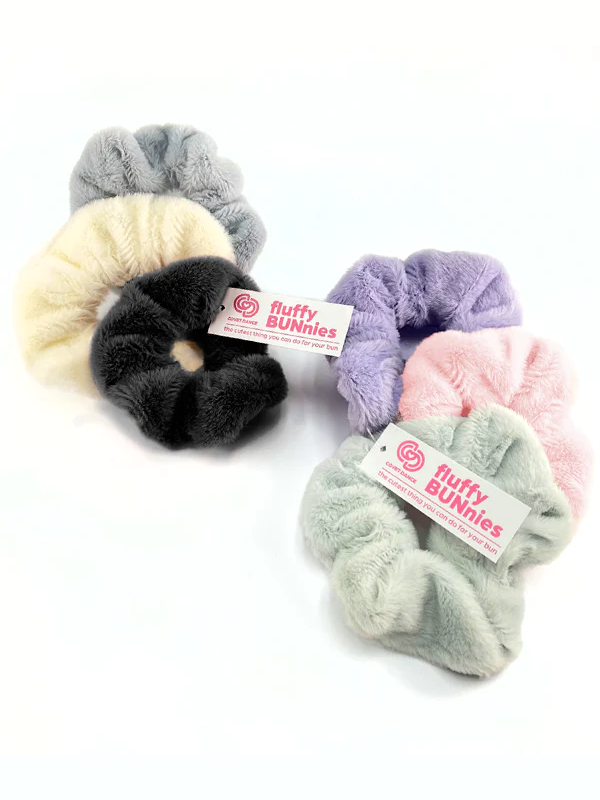 Two, three-packs of soft scrunchies next to each other. The left set contains a black, neutral white, and grey scrunchie, the right a lilac, light pink, and light mint green scrunchie, all fluffy and soft. A white tag with pink words fluffy BUNnies is visible. Plain background.