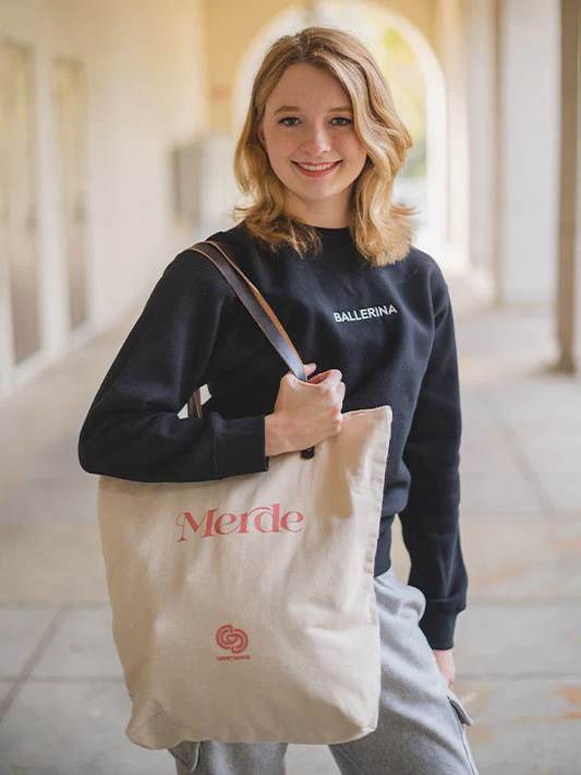 Girl Holds A Canvas Tote Bag with Leather Strap. The Word Merde is Printed On it in a Salmon Pink Color, As Well as a Logo for Covet Dance.
