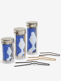 Three cylindrical containers with a blue design on the front that reads HAIRPINS and a white silhouette of a dancer's head in a bun, sit in height order: short, medium, long. Three single u-shaped hairpins sit on the ground next to each container in reverse length order, a black long pin, blonde medium pin, and short brown pin. Plain background.