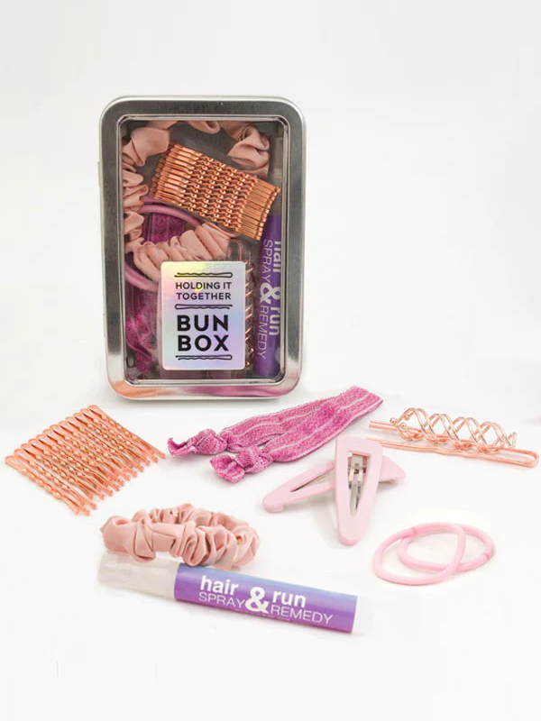 Metal tin bun box with clear lid labeled 'Holding It Together Bun Box' featuring essential ballet bun accessories: 12 rose gold bobby pins, 3 rose hair pins, 2 corkscrew spin pins, hair spray, 2 metallic pink hair ties, 2 thick pink hair elastics, 2 hair clips, and a satin scrunchie.