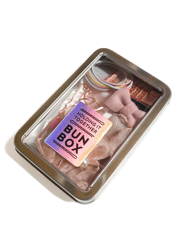 Metal tin bun box with clear lid labeled 'Holding It Together Bun Box' featuring essential ballet bun accessories: 12 rose gold bobby pins, 3 rose hair pins, 2 corkscrew spin pins, hair spray, 2 metallic pink hair ties, 2 thick pink hair elastics, 2 hair clips, and a satin scrunchie.