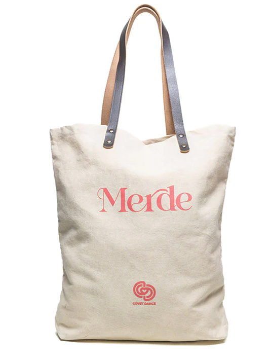 A Canvas Tote Bag with Elevated Leather Strap. The Word Merde is Printed On it in a Salmon Pink Color, As Well as a Logo for Covet Dance.