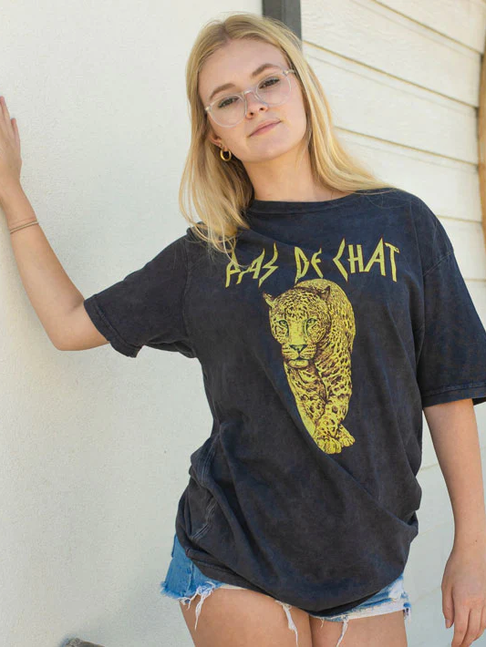 A young female with long blonde hair and clear glasses leans against a wall wearing a grey vintage inspired tee that says PAS DE CHAT and has the image of a leopard. She is wearing jean shorts with ripped edges and gold hoop earrings.