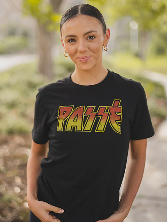 A female with pulled back hair stands outside wearing a black t-shirt with orange and yellow text that says PASSE in the style of a rock band logo, she accessorizes with gold hoop earrings.