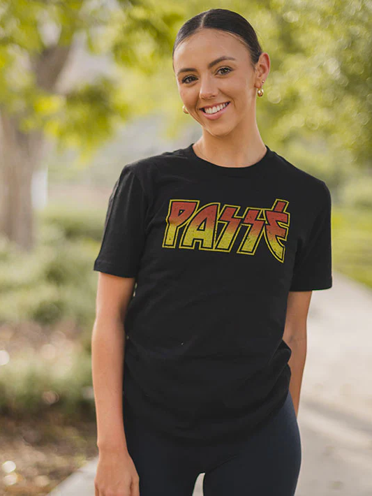 A smiling female with gold hoop earrings and pulled back hair stands outside wearing a black t-shirt with the word PASSE in a red-orange, yellow gradient.