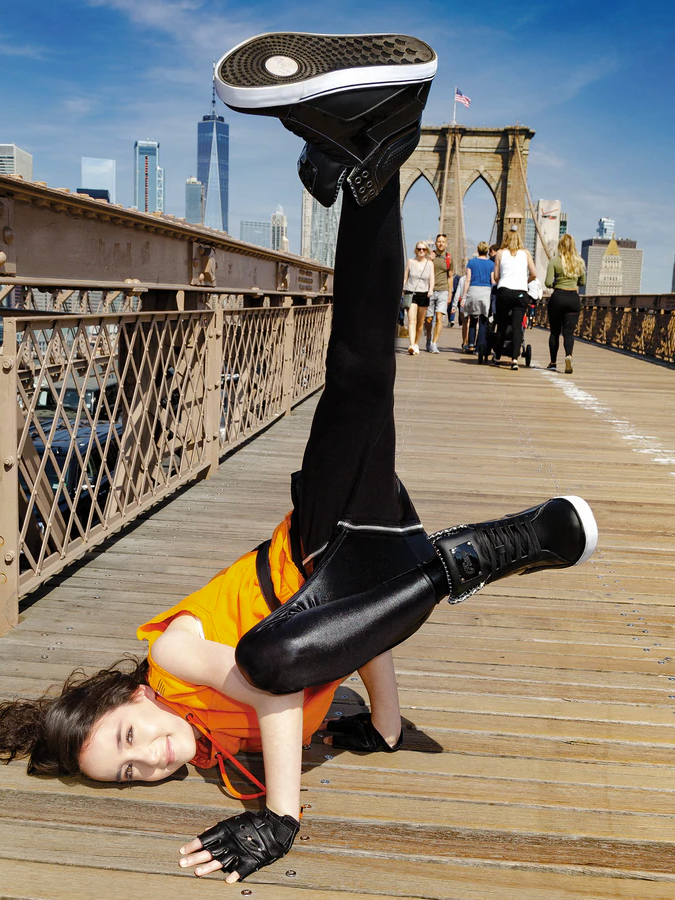 
                      
                        Female dancer wearing an orange shirt and black leggings  in break dance position on a bridge, featuring her black Pastry hip-hop sneakers
                      
                    