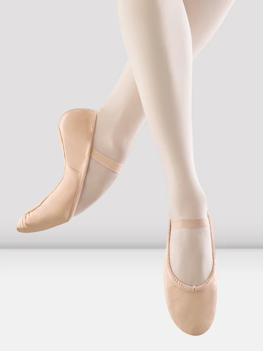 A close-up of a person's legs wearing ballet pink, leather ballet shoes with a single elastic strap across the top of the foot. The feet are pointing down as if mid-air, the back leg bent to show the flexibility of the shoe and pointe of the foot.