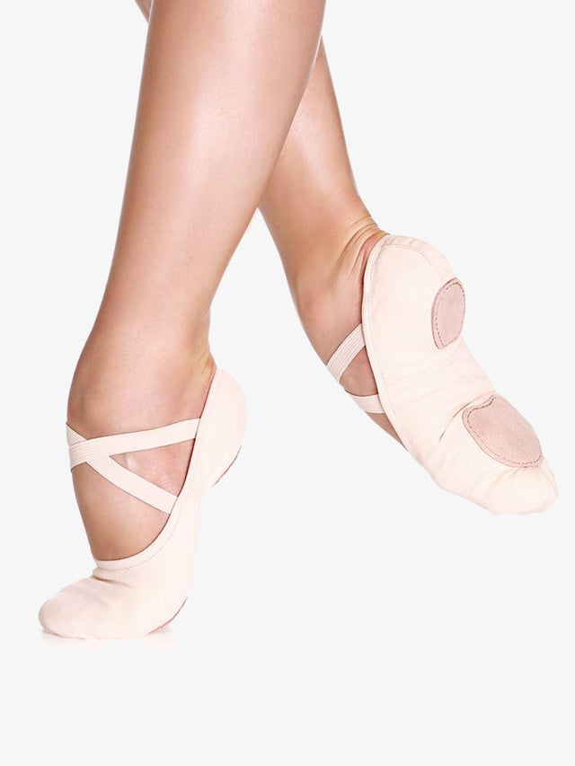 A close-up of a person's legs, wearing ballet pink, canvas ballet shoes with a criss-cross elastic design and no drawstring. The front leg is in releve and the back leg lifted and pointed to show the split-sole design and contour, against a plain background.
