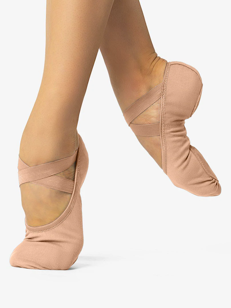 A close-up of a person's legs, wearing sand skintone, canvas ballet shoes with a criss-cross elastic design and no drawstring. The front leg is in releve and the back leg lifted and pointed to show the split-sole design and contour, against a plain background.