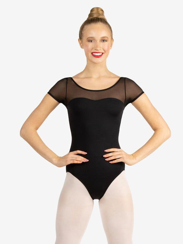 A smiling female with a blonde top bun, stands with her hands on her hips, wearing a black, mesh short sleeve, sweetheart leotard and ballet pink tights. Light grey background.