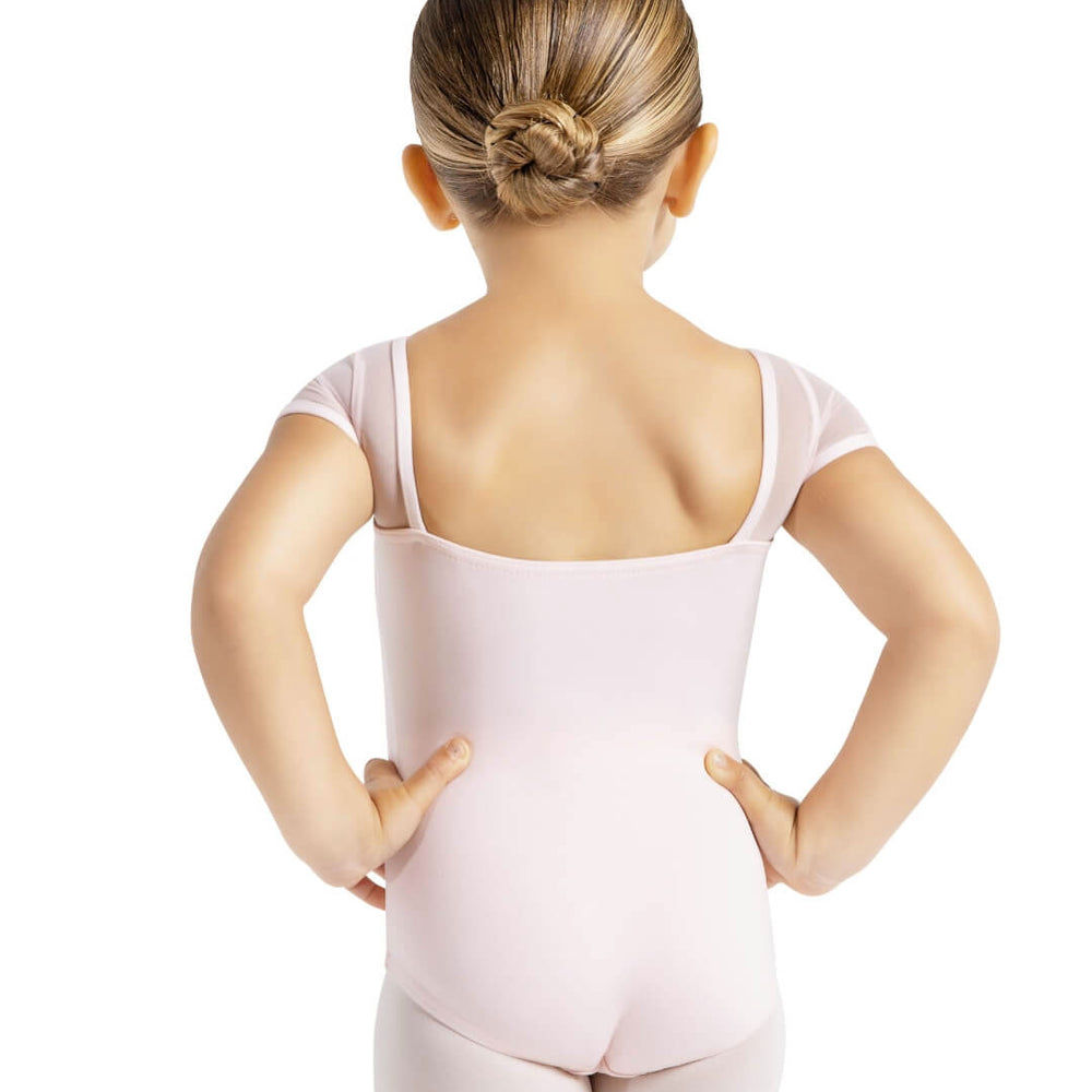 The back of a small child, hair pulled back into a ballet bun, she is wearing a light pink leotard and tights and stands with her hands on her hips.