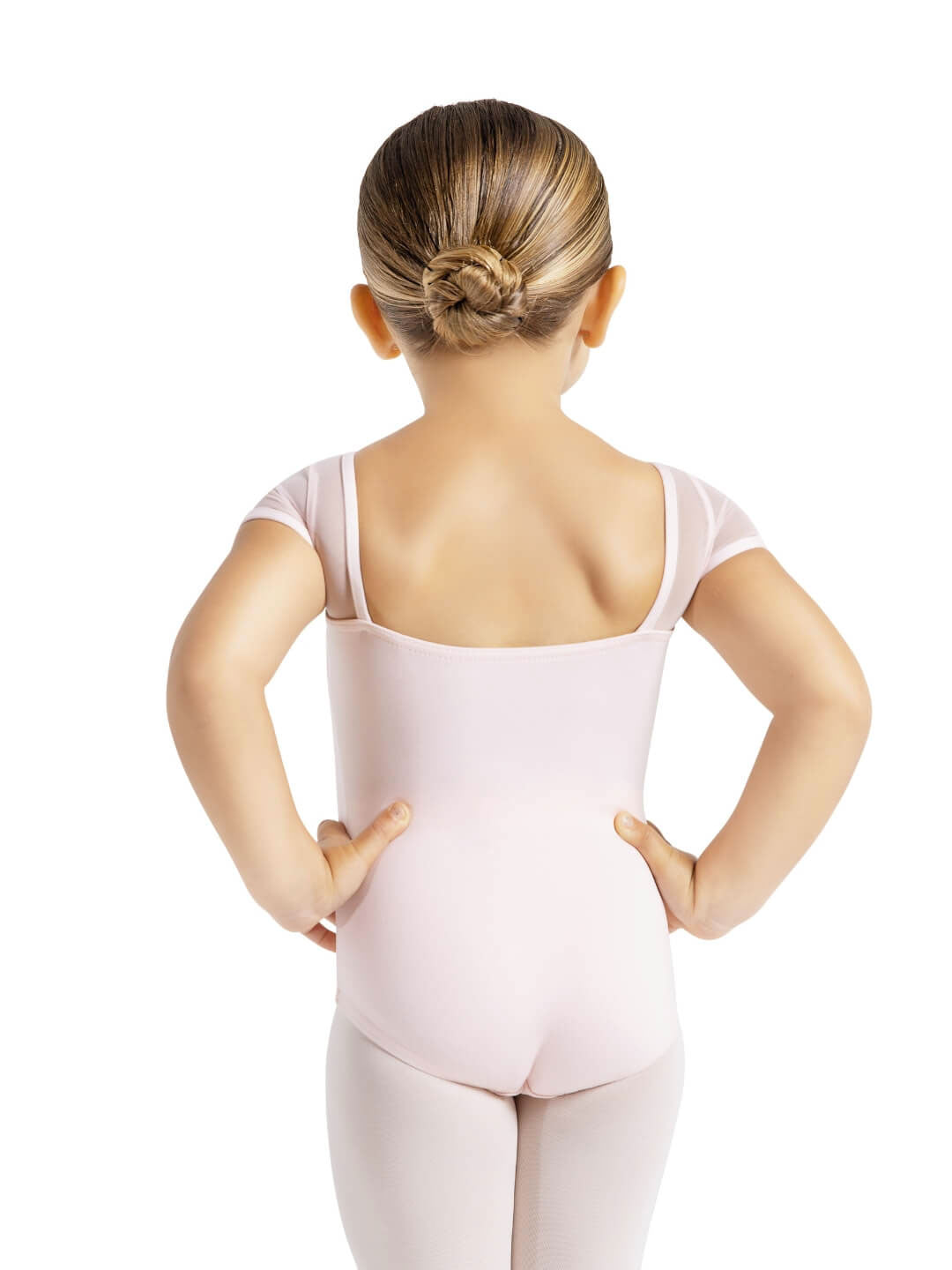 The back of a small child, hair pulled back into a ballet bun, she is wearing a light pink leotard and tights and stands with her hands on her hips.