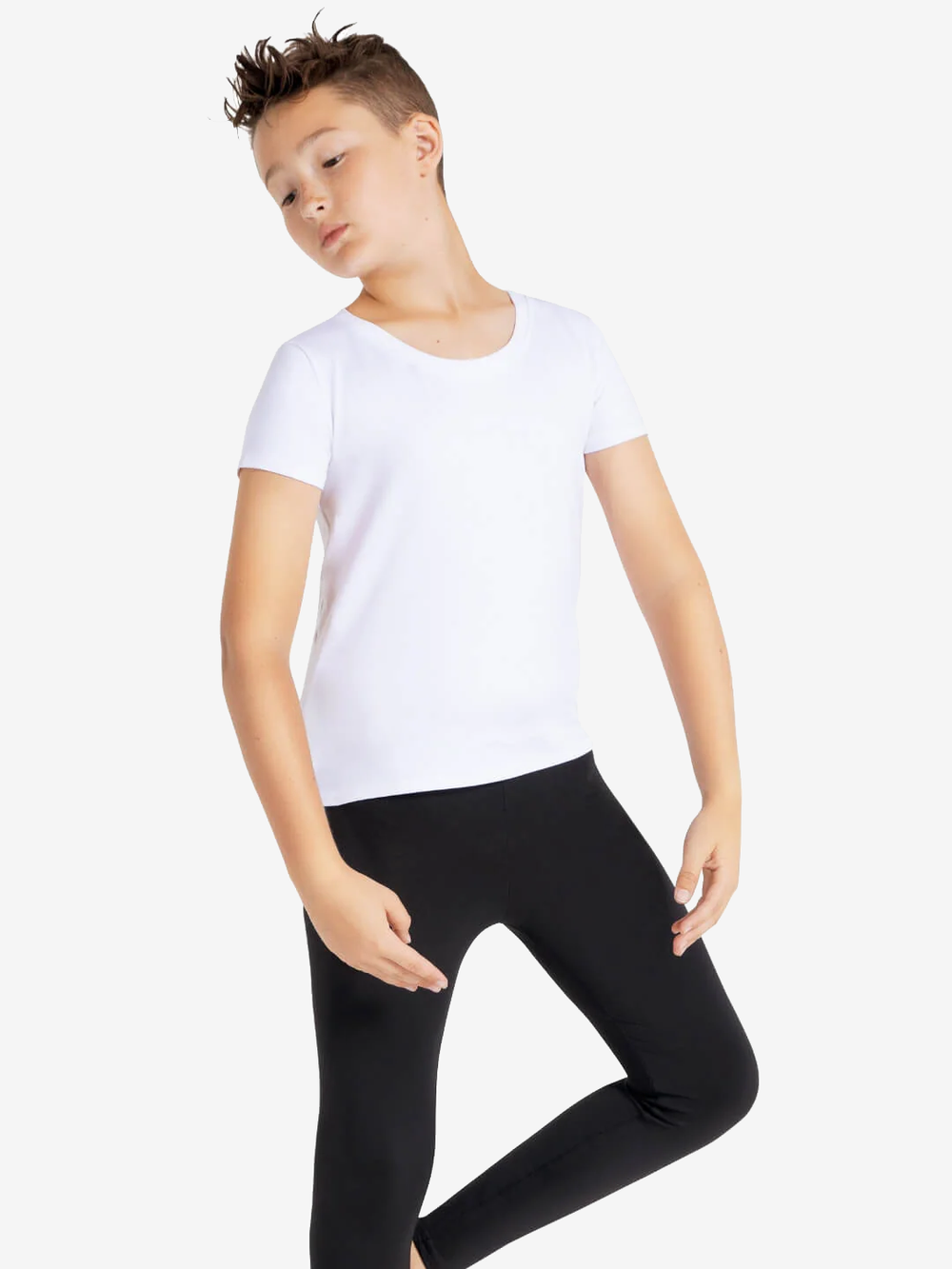 A boy with spiky hair leans back in a ballet pose wearing a white crew neck t-shirt and black leggings, against a plain light grey background.