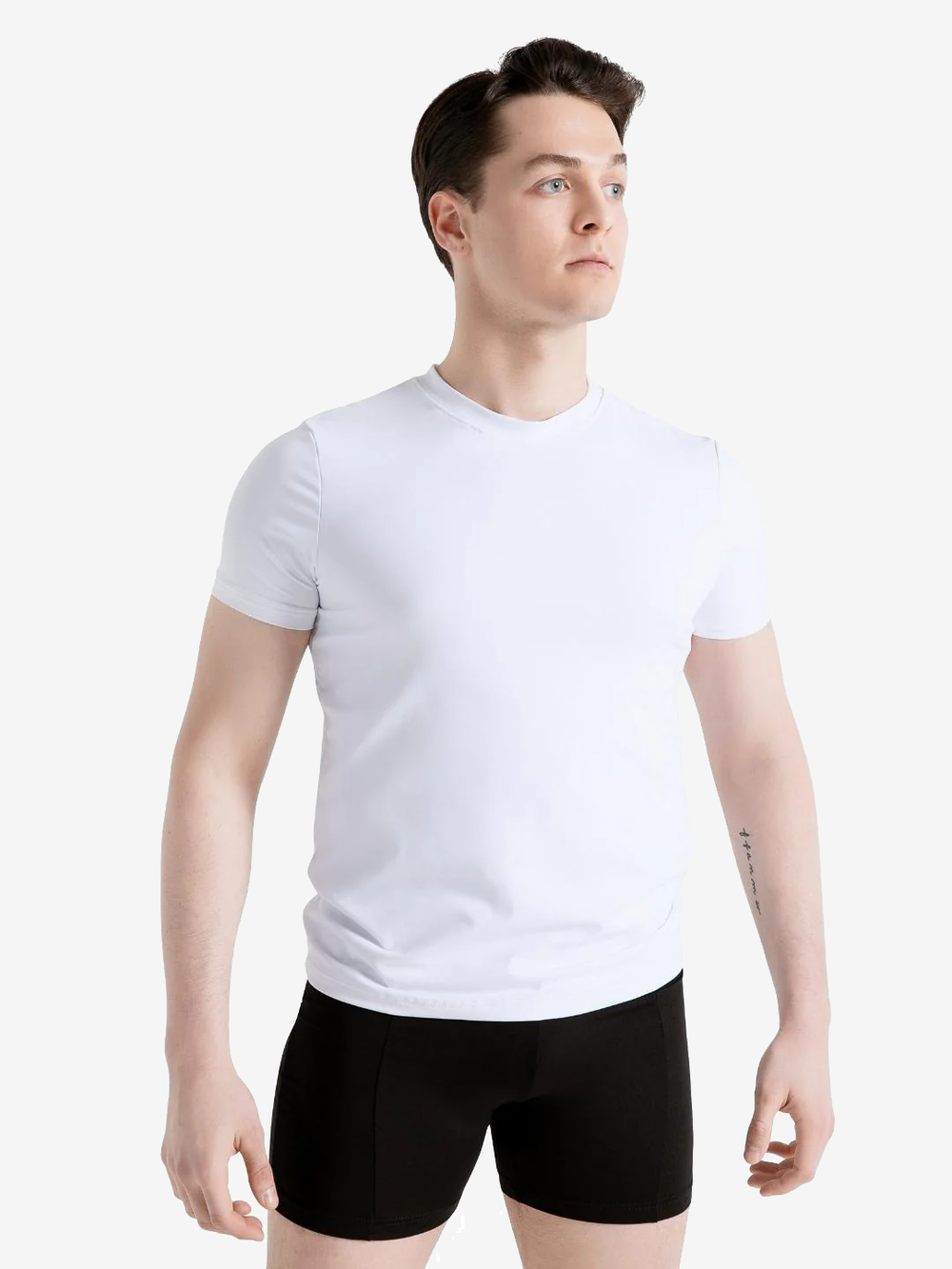 A mid-shot male with short brown hair stands arms at sides, gazing off to the side. He is wearing a white crew neck t-shirt and black dance shorts. The background is plain light grey.