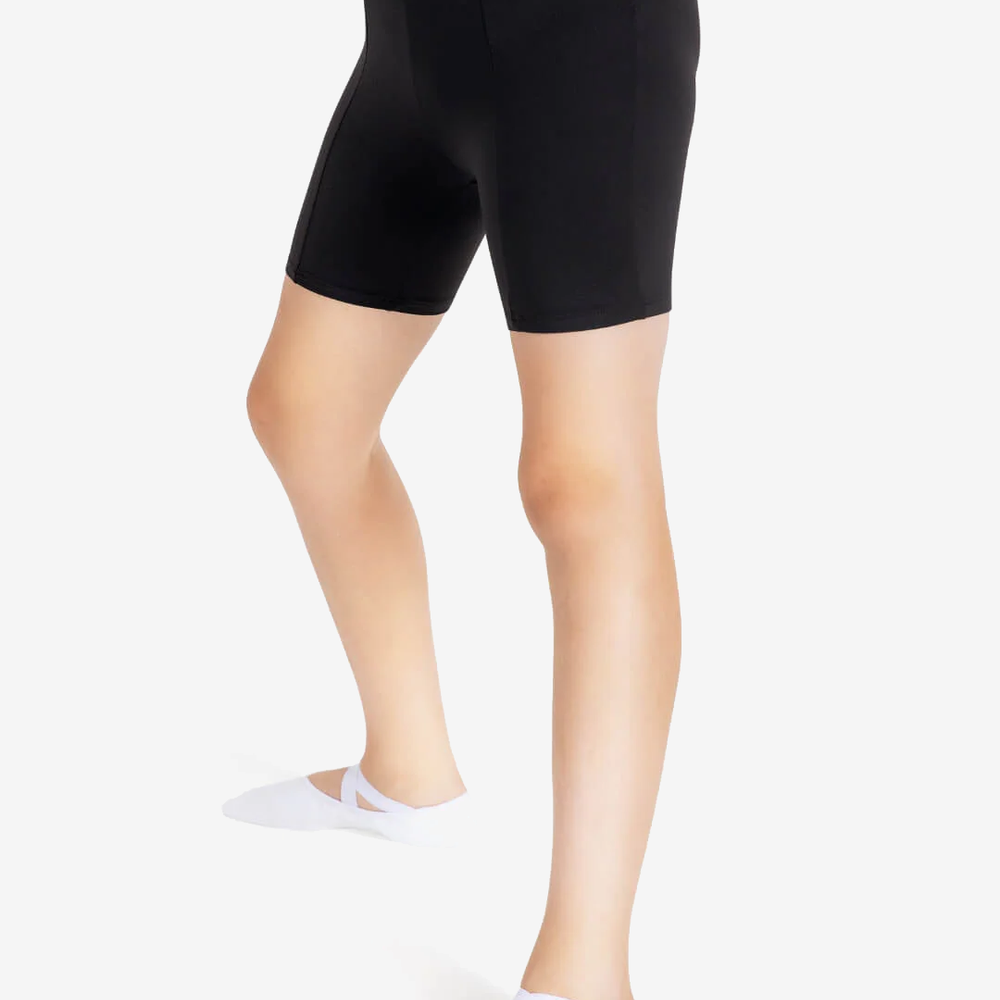 
                      
                        A close-up of a person's legs wearing black dance shorts and white ballet shoes with a criss-cross elastic, against a plain light grey background.
                      
                    