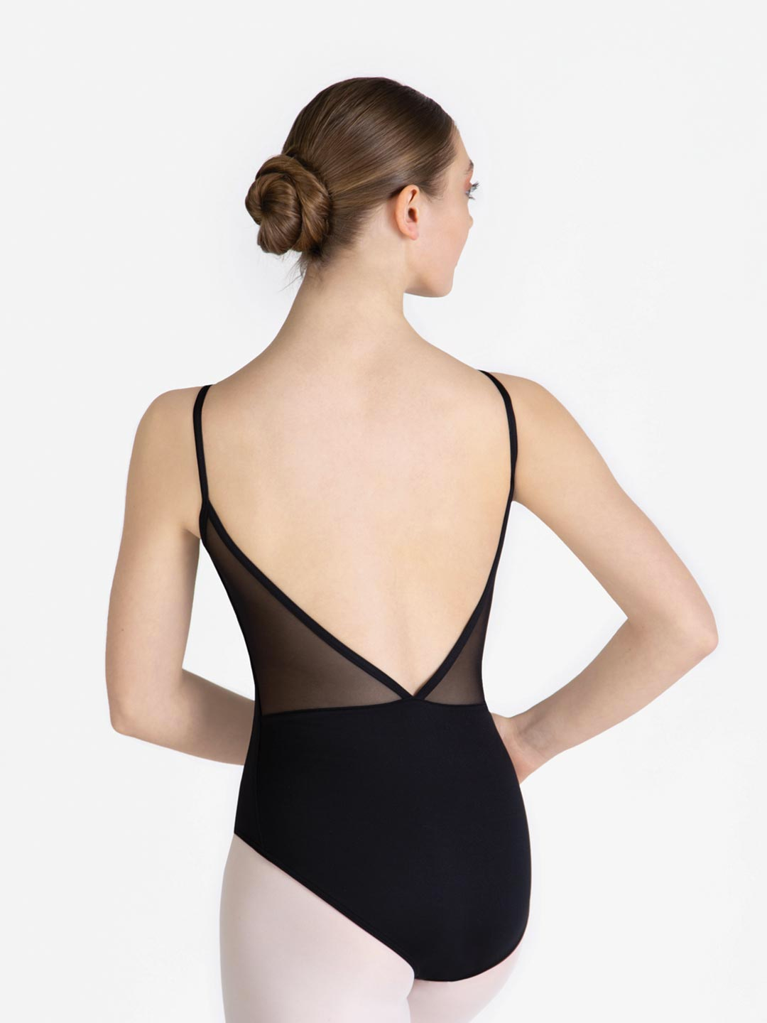 A female standing with her back facing us, hand on hips, wearing a black leotard with a mesh v-shaped low back, hair in a ballet bun.