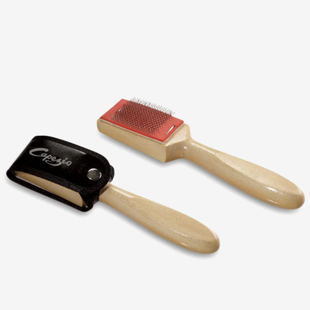 A pair of wire brushes lay side-by-side. The handles are a natural wood look, the brush is wire, one has a black cap closed over the wire brush with a snap and features a Capezio logo. Light grey background.