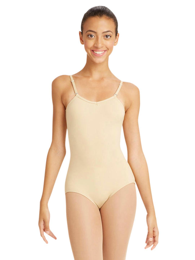 A female dancer stands against a solid white background wearing a nude colored, spaghetti strap leotard with a ballet leg line, hands held at her sides. She has dark hair pulled back.