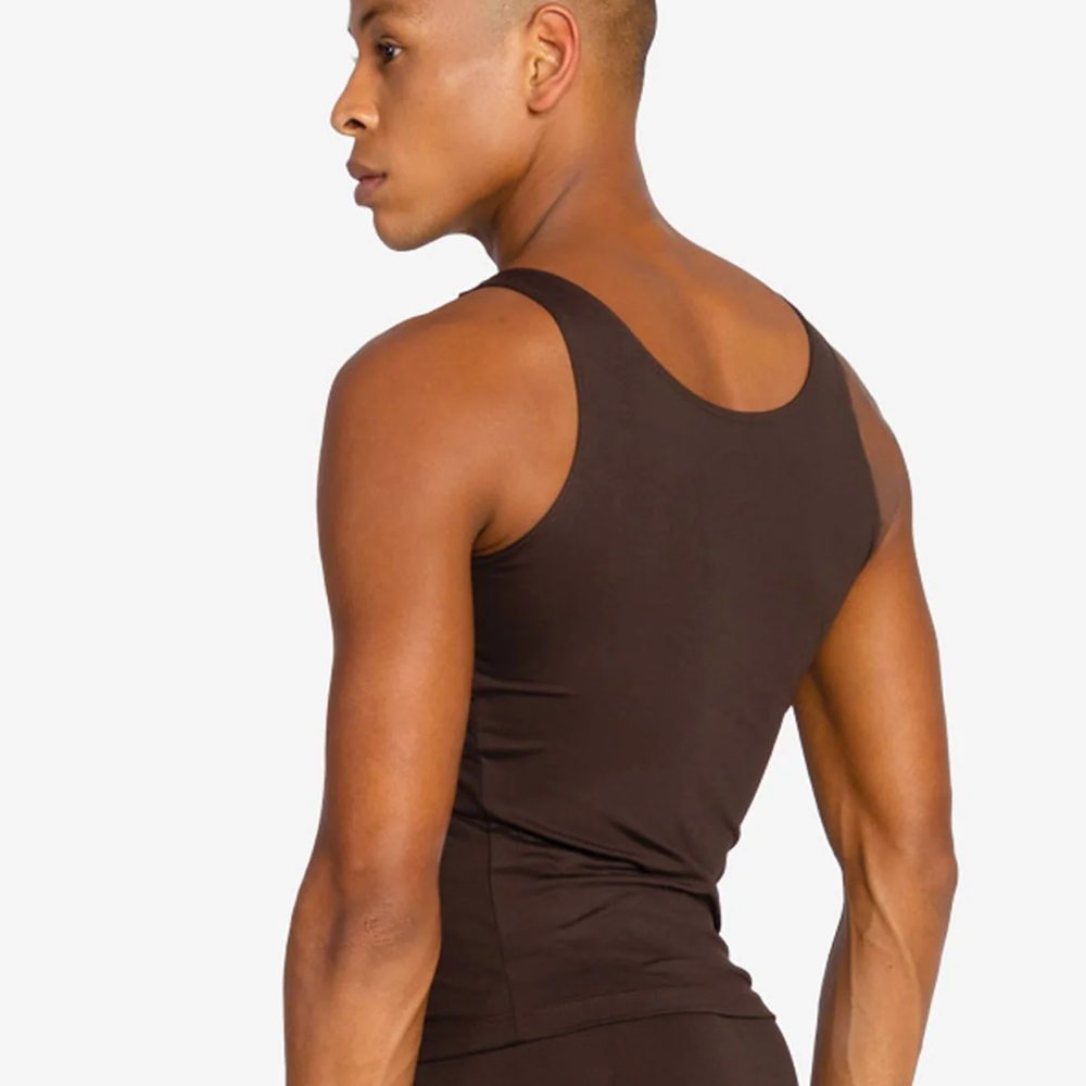 A man with dark hair on top, shaved on the sides, is turned to the side to show the back of a coffee colored tank top and ballet leggings.