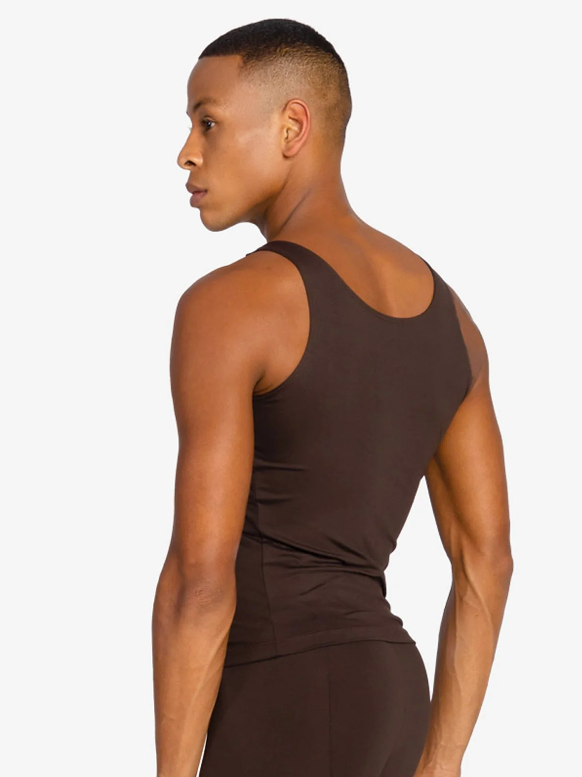 A man with dark hair on top, shaved on the sides, is turned to the side to show the back of a coffee colored tank top and ballet leggings.
