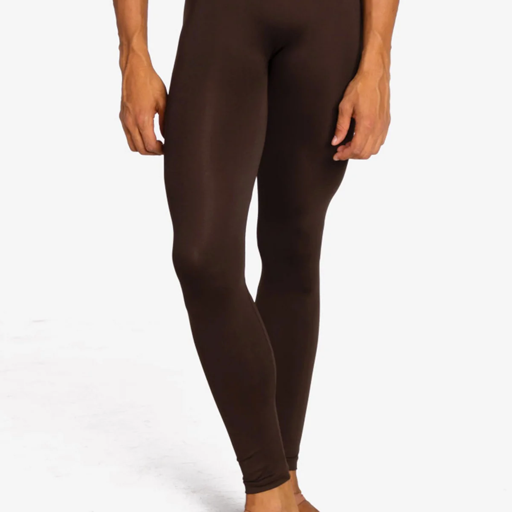 A close-up of a male dancer from the waist down wearing coffee colored tight leggings and nude canvas ballet shoes against a plain background.