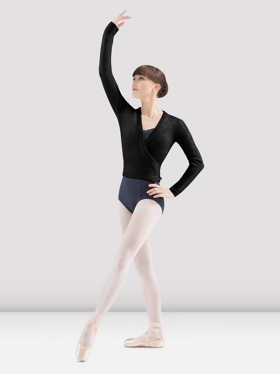 A ballet dancer in a black wrap sweater, black leotard, and pink tights poses gracefully. She stands in pointe shoes with one arm elevated and the other hand resting on her hip. The background is plain grey.