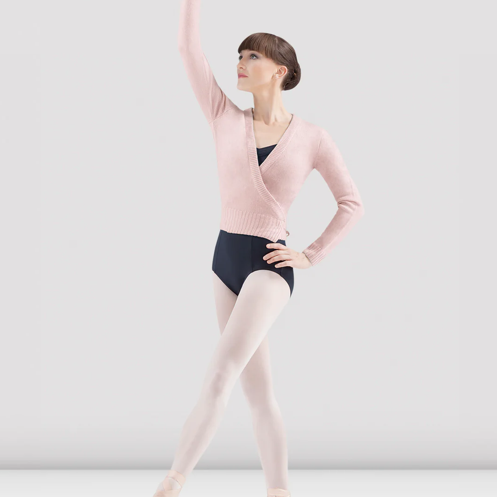 A ballet dancer in a light pink wrap sweater, black leotard, and pink tights poses gracefully. She stands in pointe shoes with one arm elevated and the other hand resting on her hip. The background is plain grey.
