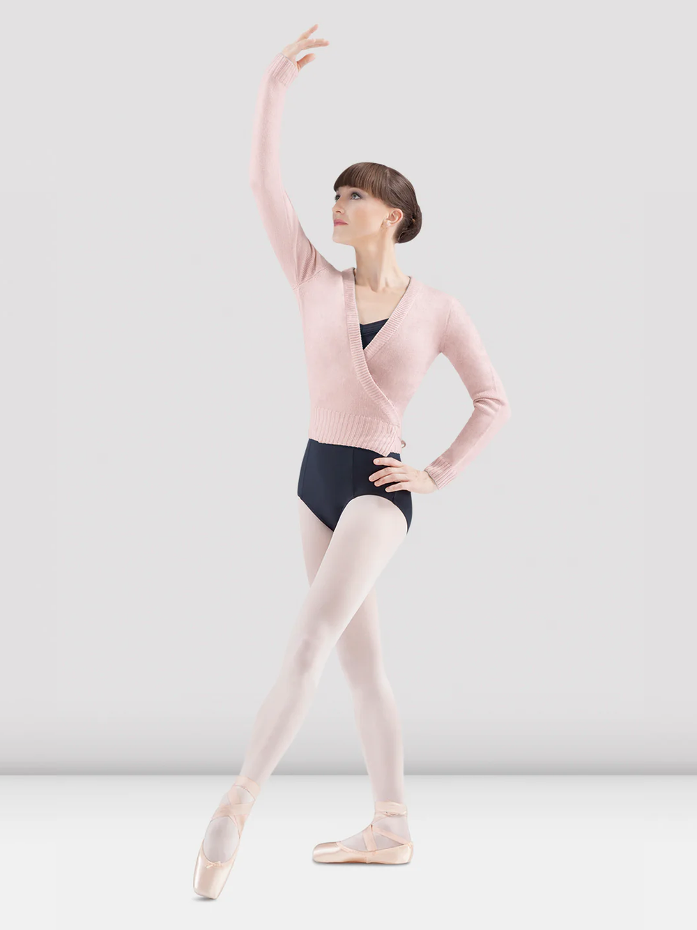 A ballet dancer in a light pink wrap sweater, black leotard, and pink tights poses gracefully. She stands in pointe shoes with one arm elevated and the other hand resting on her hip. The background is plain grey.