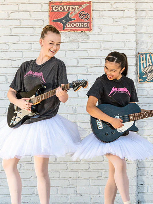 Two young girls in tutus playing electric guitars against a white brick wall with 'Covet Rocks' sign.