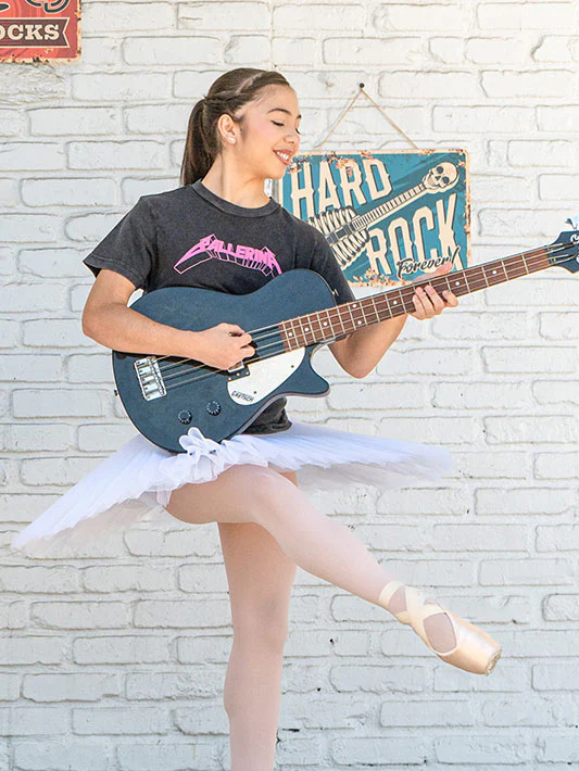 
                      
                        Girl in ballet outfit playing a blue bass guitar against a white brick wall with a Hard Rock Cafe sign.
                      
                    