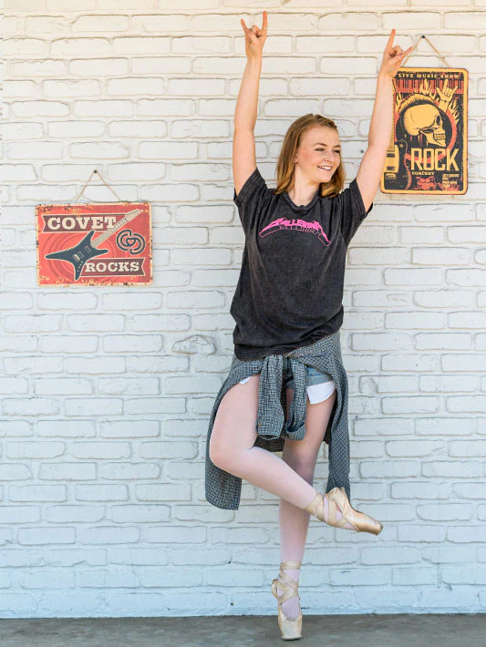 
                      
                        Girl in a black t-shirt with text, gray skirt, and beige shoes posing against a white brick wall with posters.
                      
                    