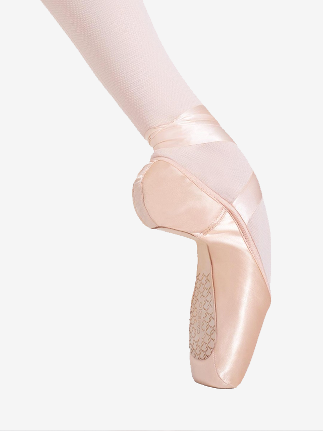 A close-up of a person's single foot extended and en pointe wearing Capezio Cambre and pink tights with ribbon tied up the ankle, showcasing the arch and flexibility in the shank of the shoe.