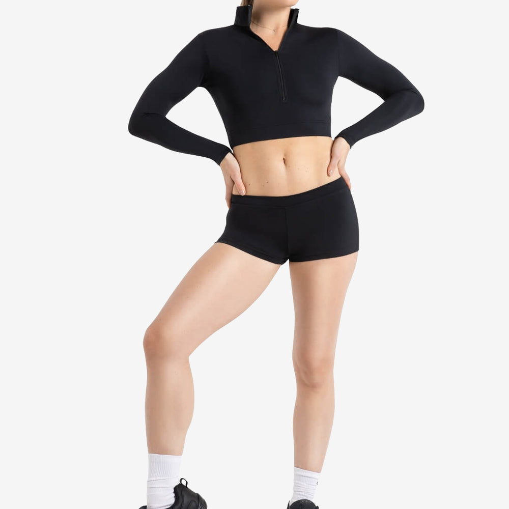 
                      
                        A person stands, hands on hip, wearing a zip-up, black, long sleeve crop top and fitted black shorts, white socks, and black dance sneakers. One foot is pointed down to show pointe and flexibility. Light grey background.
                      
                    