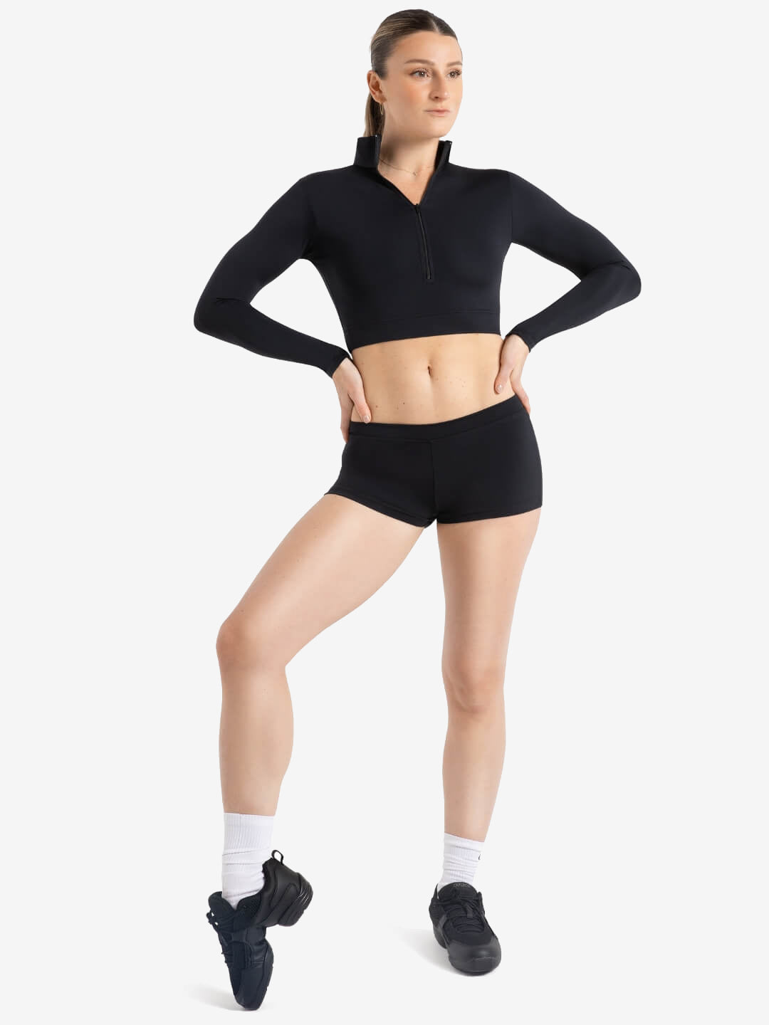 A person stands, hands on hip, wearing a zip-up, black, long sleeve crop top and fitted black shorts, white socks, and black dance sneakers. One foot is pointed down to show pointe and flexibility. Light grey background.