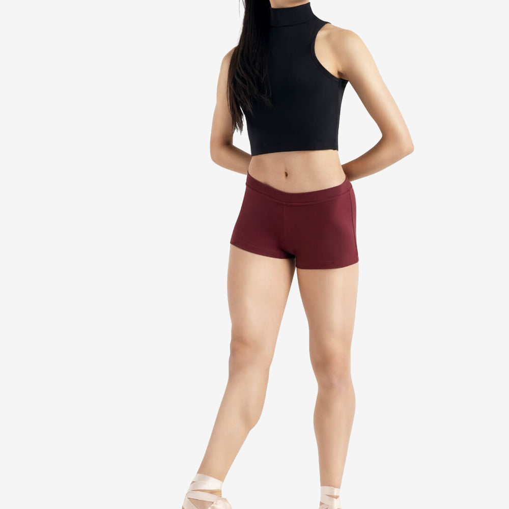 
                      
                        A person wearing a black sleeveless top, maroon shorts, and pointe shoes stands in a relaxed ballet pose with one leg extended and hands behind their back, set against a plain light grey background.
                      
                    