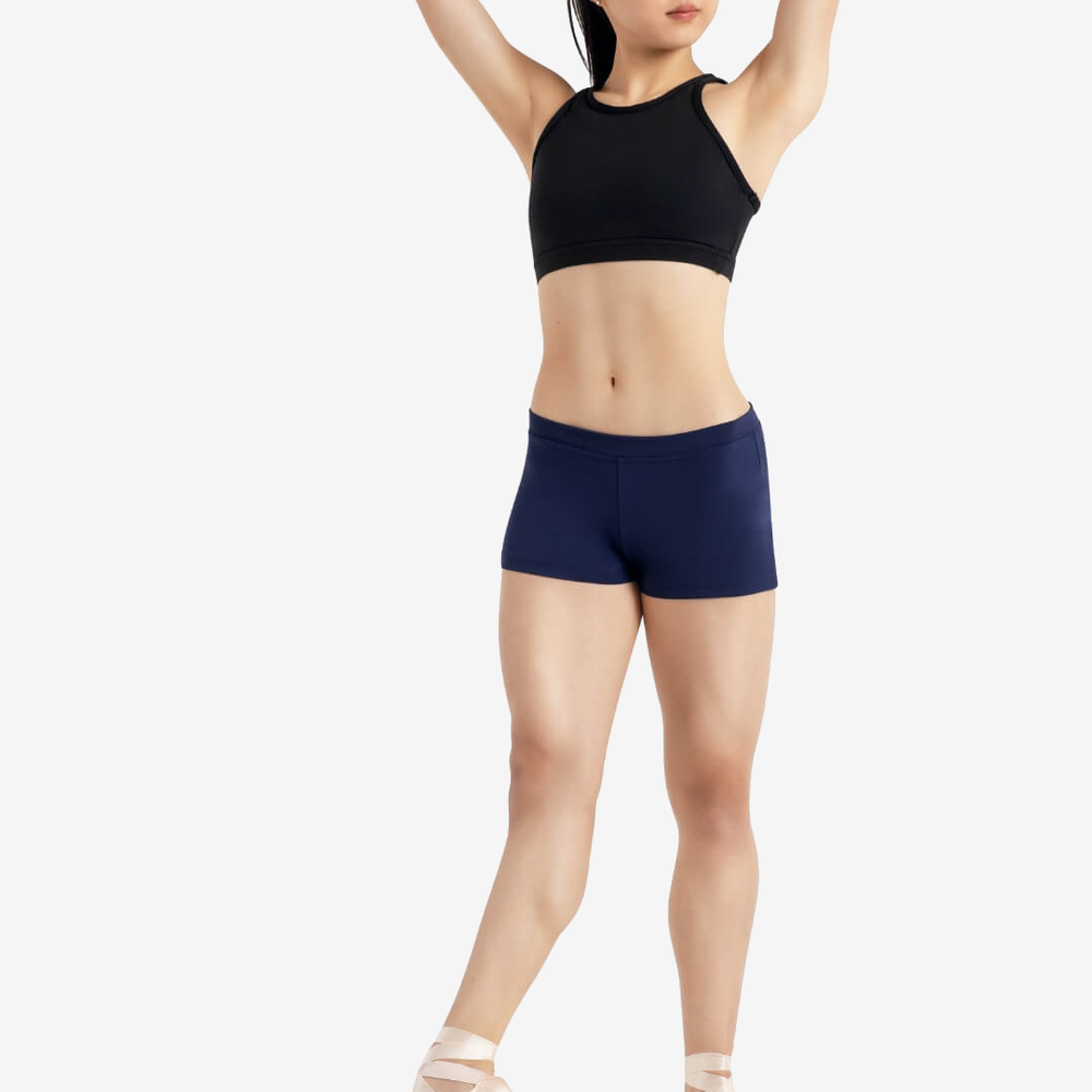 
                      
                        A person wearing a black sleeveless top, navy shorts, and pointe shoes stands in a relaxed ballet pose with one leg extended and hands behind their head, set against a plain light grey background.
                      
                    