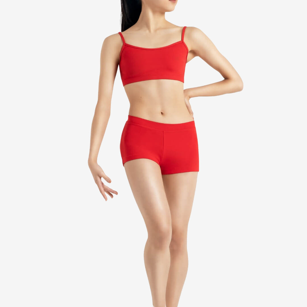 
                      
                        A person wearing a red sports bra, red shorts, and pointe shoes stands in a relaxed ballet pose with one leg slightly bent and one hand on her hip, the other at her side, set against a plain light grey background.
                      
                    