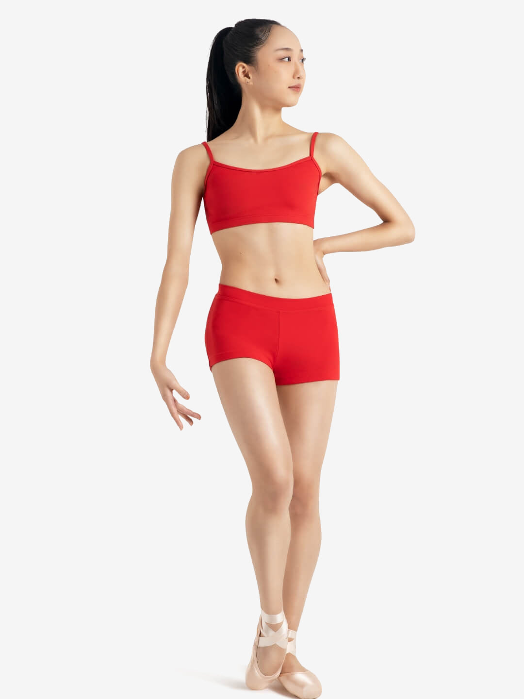 A person wearing a red sports bra, red shorts, and pointe shoes stands in a relaxed ballet pose with one leg slightly bent and one hand on her hip, the other at her side, set against a plain light grey background.