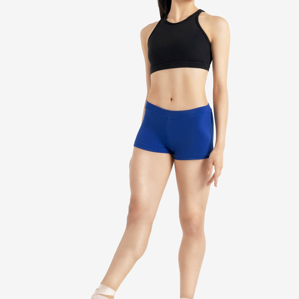 
                      
                        A person wearing a black sleeveless top, royal blue shorts, and pointe shoes stands in a relaxed ballet pose with one leg extended and hands at their sides, set against a plain light grey background.
                      
                    