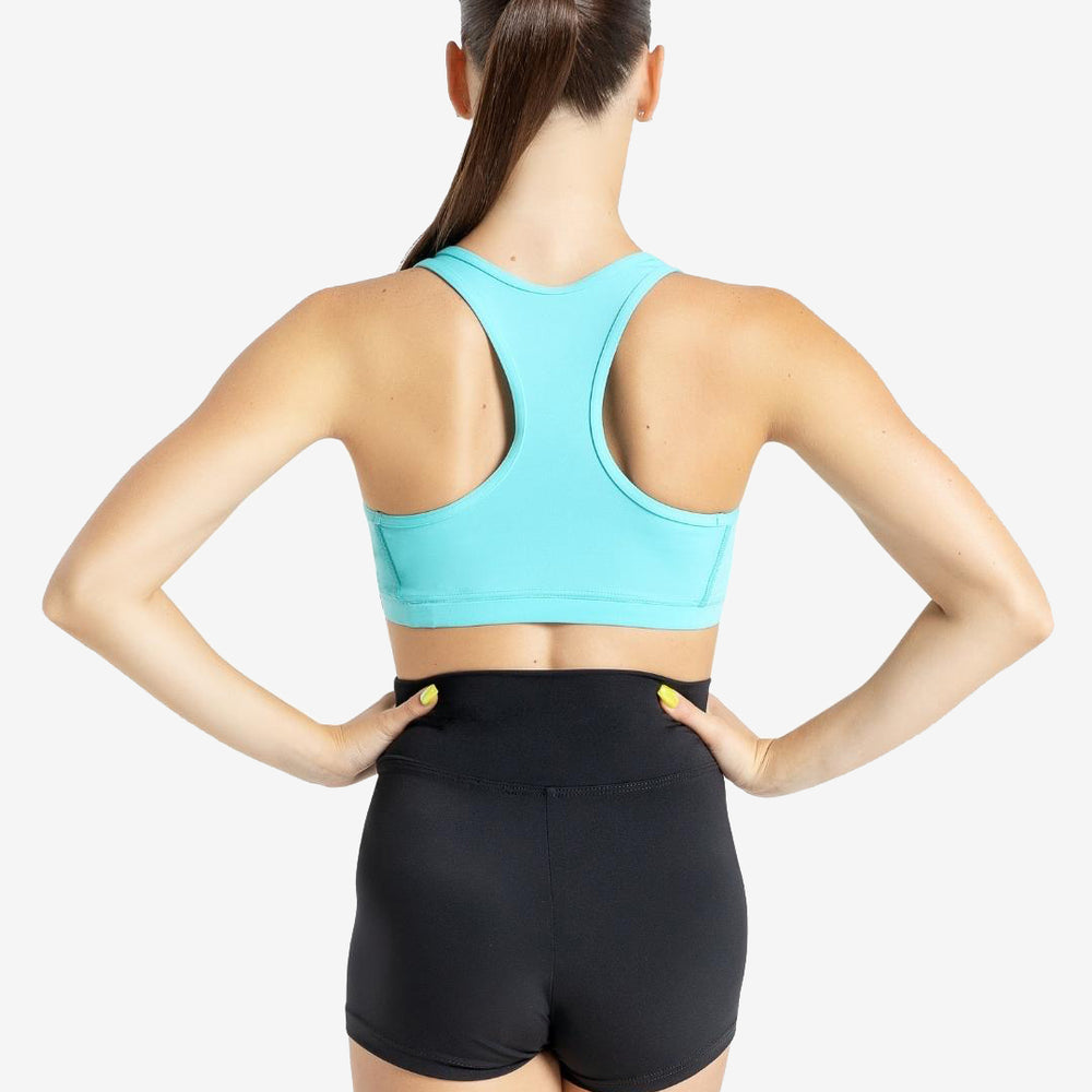 The back of a woman with a long brown ponytail draped over her front shoulder wearing a turquoise sports bra and high waisted black shorts. Her hands are on her hips with painted yellow nails.